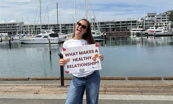 Photo of a woman standing by Wellington waterfront with a sign saying What makes a healthy relationship?